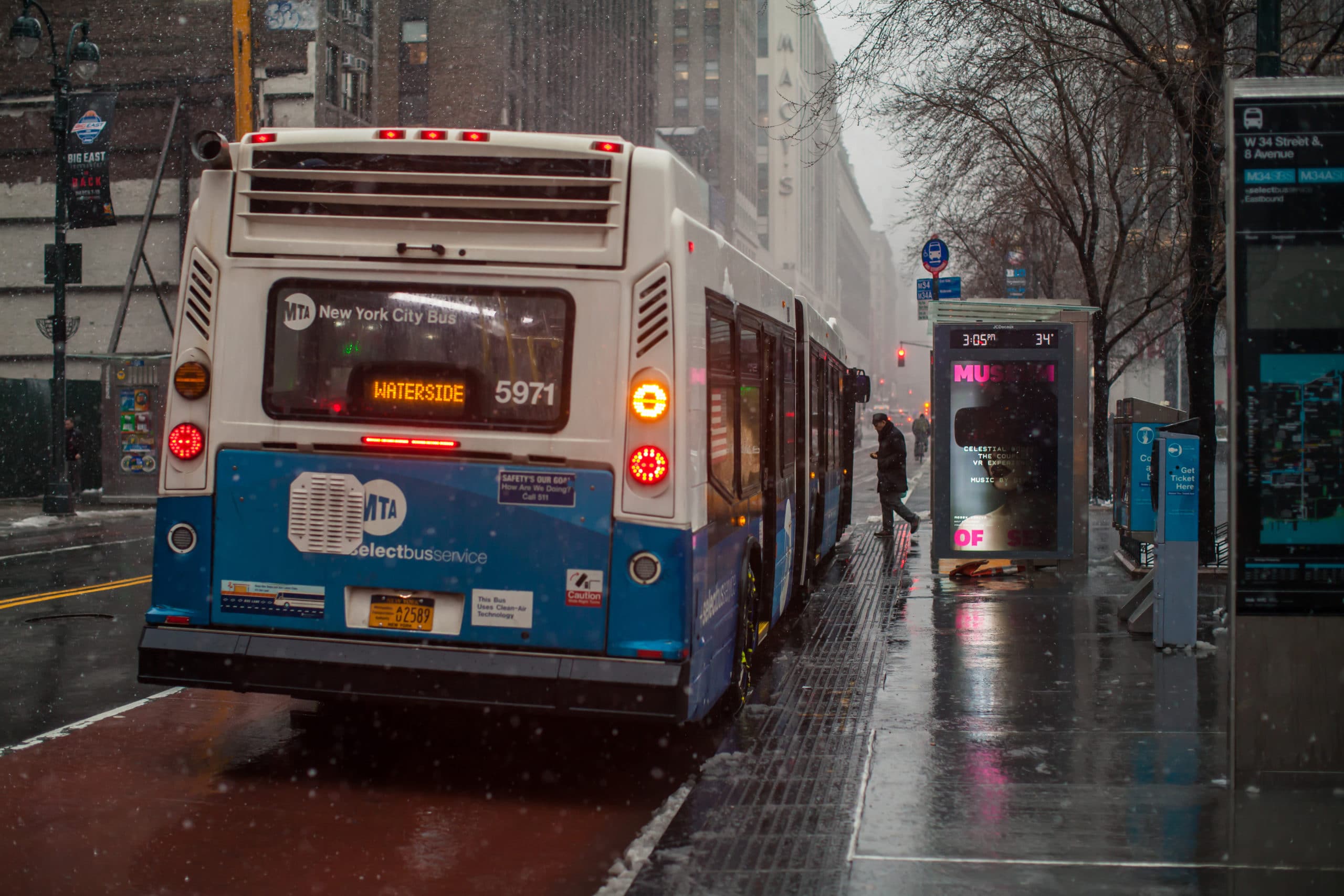 New York City MTA bus at a bus stop. A man in a coat enters the front door of the bus. The roadway is wet and glossy. Traffic lights, trucks and cars visible on the photo. High-rise buildings on the left and right. In the distance, skyscrapers are in snowy haze. Snow continues to fall and snowflakes are visible flying in the air. Penn station area. Slippery. Very bad weather. Evening. March 07, 2018. NYC. Midtown Manhattan. New York. USA